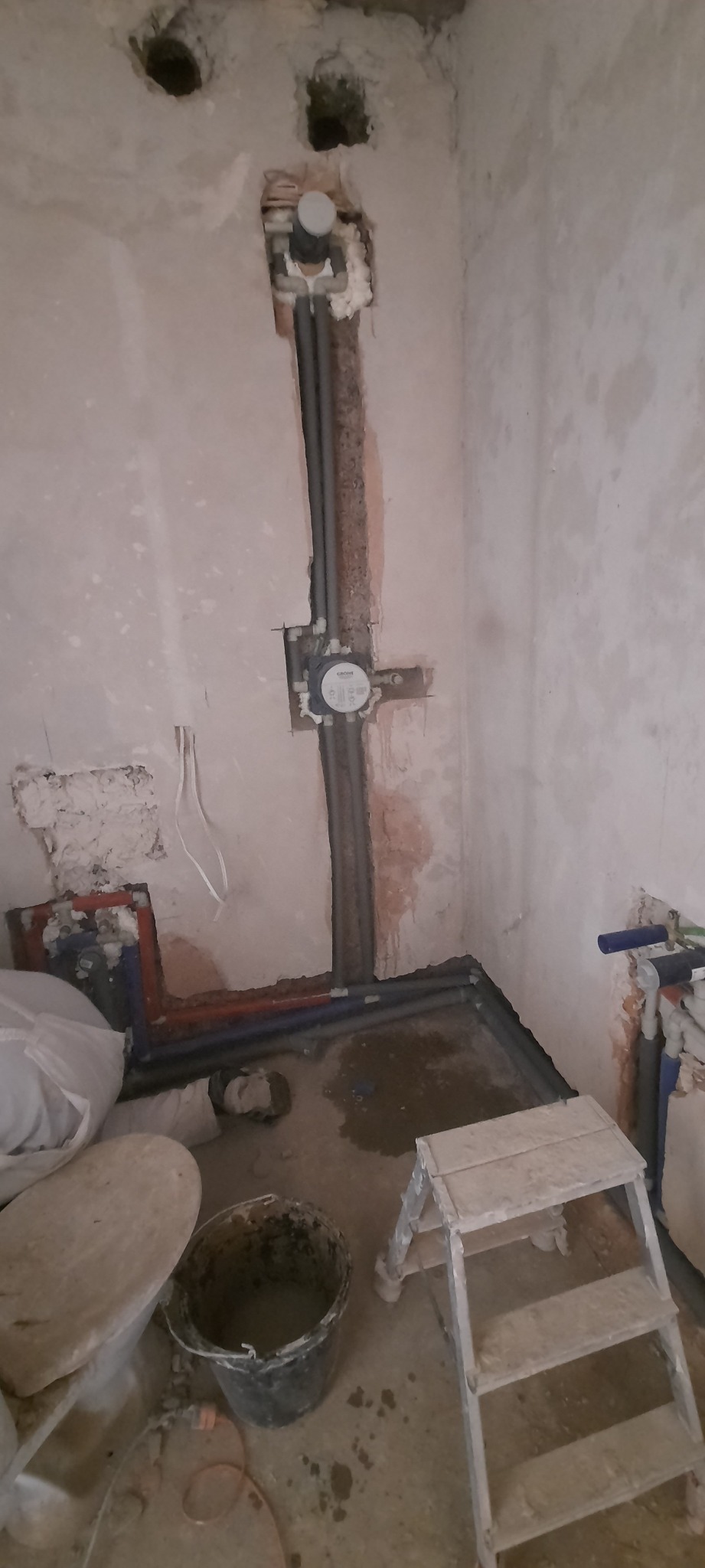 Surowe ściany w trakcie prac instalacyjnych w łazience, widoczne rury, puszka podtynkową i drabina malarska.