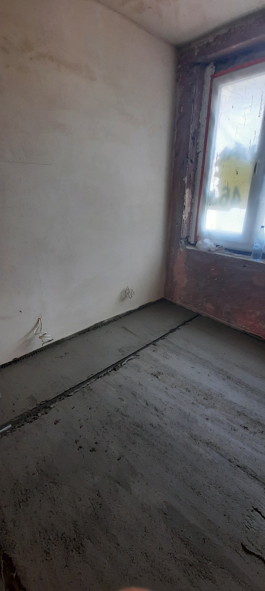 Surowy, szary beton na podłodze w trakcie wylewania, widoczne listwy dylatacyjne, ściany w stanie surowym, okno zabezpieczone folią malarską.
