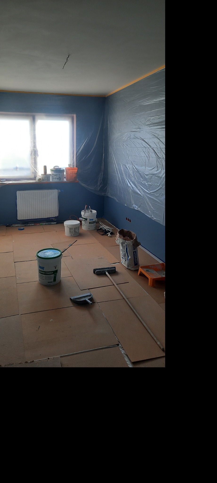 Przygotowania do prac wykończeniowych: zabezpieczona folią ściana pomalowana na niebiesko, karton na podłodze, wiadro z gładzią Knauf Super Finish, pędzel i szpachelka