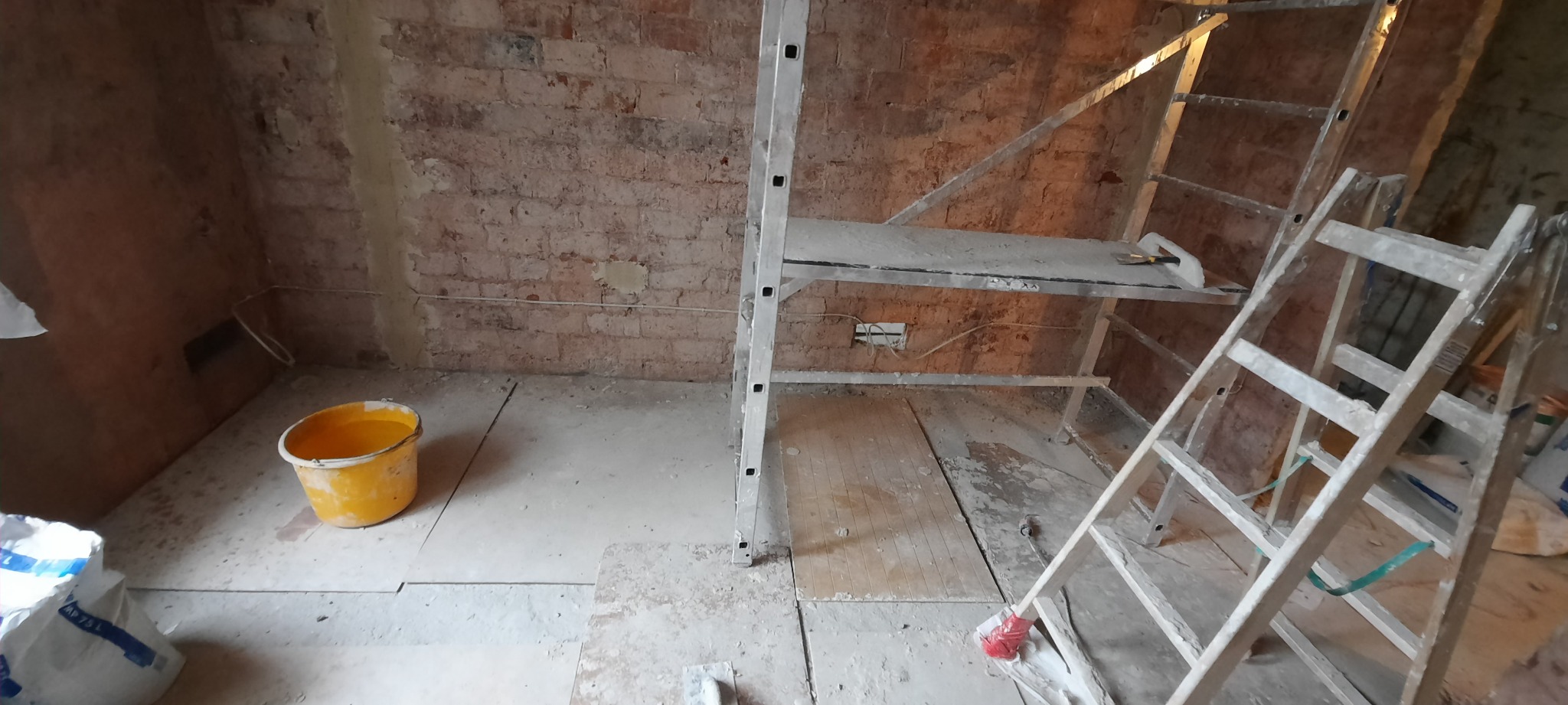 Wnętrze w trakcie prac przygotowawczych: surowa cegła, częściowo zagruntowana ściana, drabiny, rusztowanie i wiadro z płynem.
