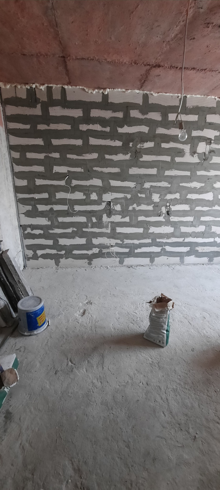 Murowana ściana z betonu komórkowego w trakcie prac wykończeniowych, widoczne fugi wypełnione zaprawą, surowy betonowy strop, wiadro z farbą i worek z zaprawą na podłodze.
