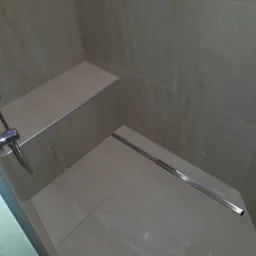 Wykończona kabina prysznicowa z siedziskiem i odpływem liniowym, wyłożona jasnymi płytkami imitującymi kamień, widoczna chromowana bateria.