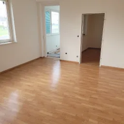 Pusty pok&oacute;j z panelami podłogowymi w odcieniach brązu, świeżo pomalowanymi ścianami na biało i otwartymi drzwiami do kolejnych pomieszczeń, w kt&oacute;rych widać materiały budowlane.