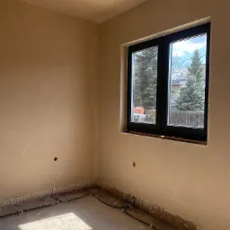 Wnętrze pokoju po nałożeniu tynków, widoczne ślady po instalacji elektrycznej, czarne ramy okienne z widokiem na iglaki na zewnątrz.