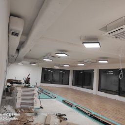 Lux Max - Wnętrze w trakcie montażu oświetlenia i klimatyzacji: widoczne kanały wentylacyjne, klimakonwektory kasetonowe, panele LED i częściowo ułożona podłoga z paneli drewnopodobnych.