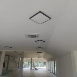 Wnętrze w trakcie wykańczania: widok na sufit z zainstalowanymi kwadratowymi lampami LED z czarną obwódką i klimatyzatorem kasetonowym, widoczne również filary i okna z widokiem na zieleń.