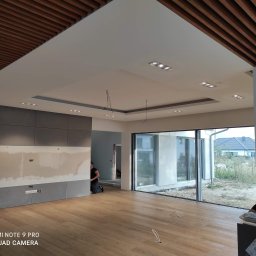 Lux Max - Wnętrze w trakcie wykańczania: drewniana podłoga, szare panele ścienne z widocznymi przewodami elektrycznymi, sufit z drewnianymi listwami i zamontowanymi punktowymi oprawami oświetleniowymi, osoba...