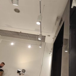 Lux Max - Montaż szynoprzewodów oświetleniowych z reflektorami LED w pomieszczeniu z kratką wentylacyjną na suficie, widoczny fragment sylwetki montera i lampy warsztatowej.