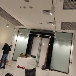 Lux Max - Elektryk instaluje oświetlenie szynowe w nowym lokalu, widoczne narzędzia i materiały przygotowane do montażu, w tle szklane ściany i białe drzwi.