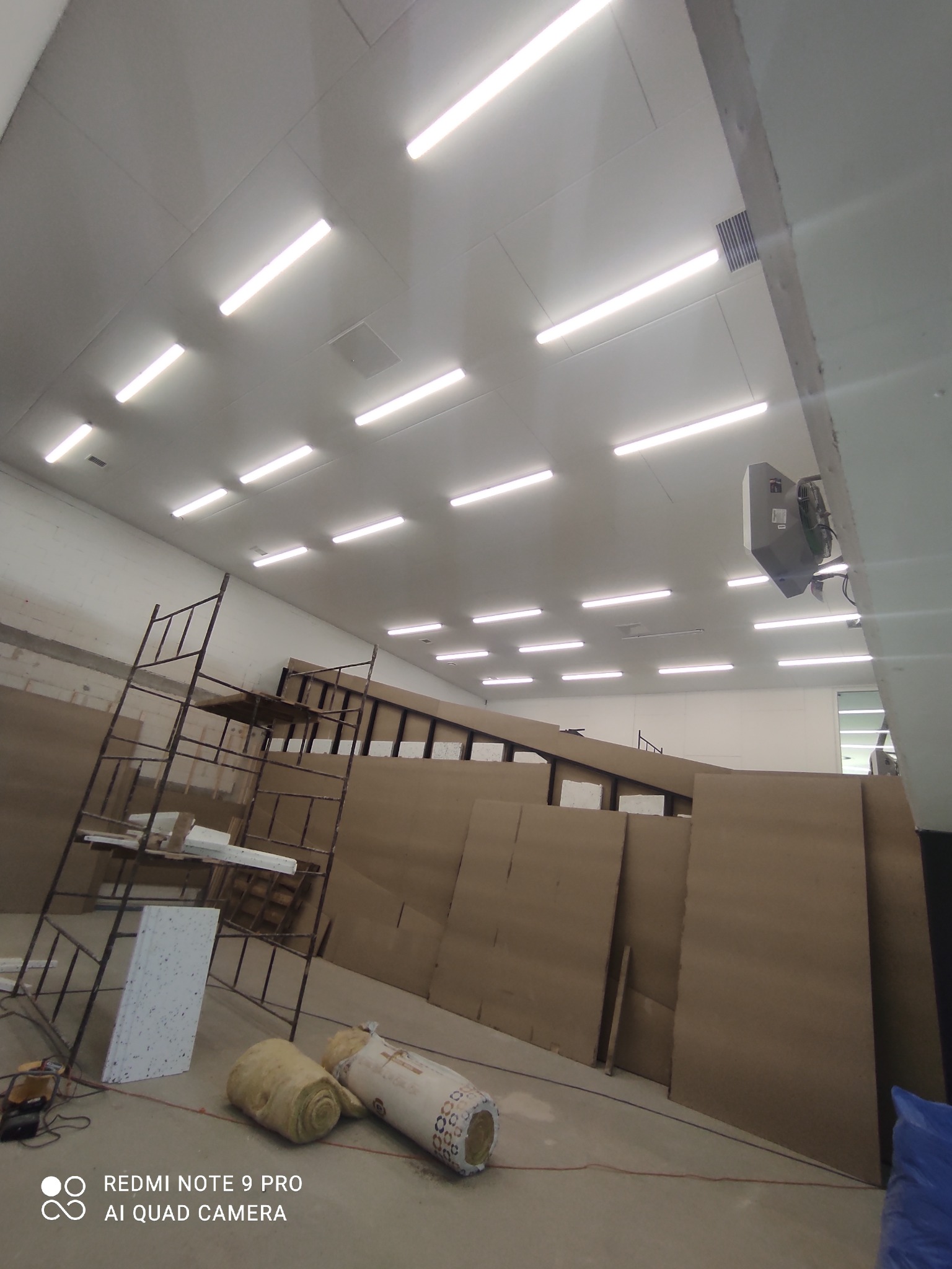 Montaż oświetlenia LED w hali przemysłowej: widoczne lampy, rusztowanie, materiały izolacyjne i wentylator przemysłowy na ścianie.