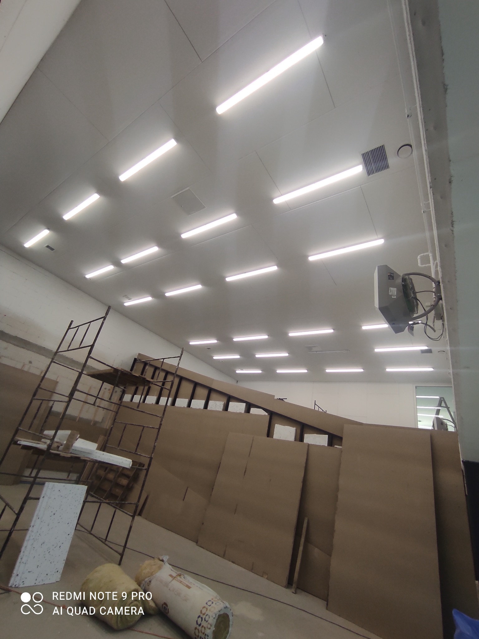 Montaż oświetlenia sufitowego w dużej hali, widoczne panele sufitowe, lampy LED, rusztowania i materiały izolacyjne w rolkach.