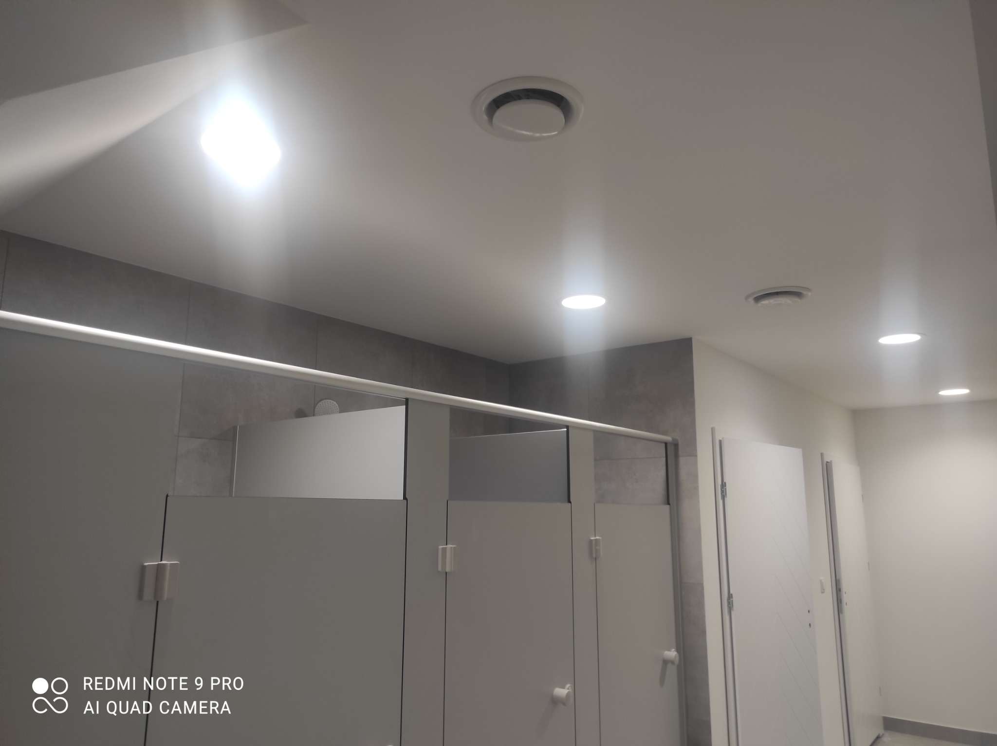 Wnętrze publicznej toalety z kabinami, widoczne oświetlenie sufitowe i kratki wentylacyjne.