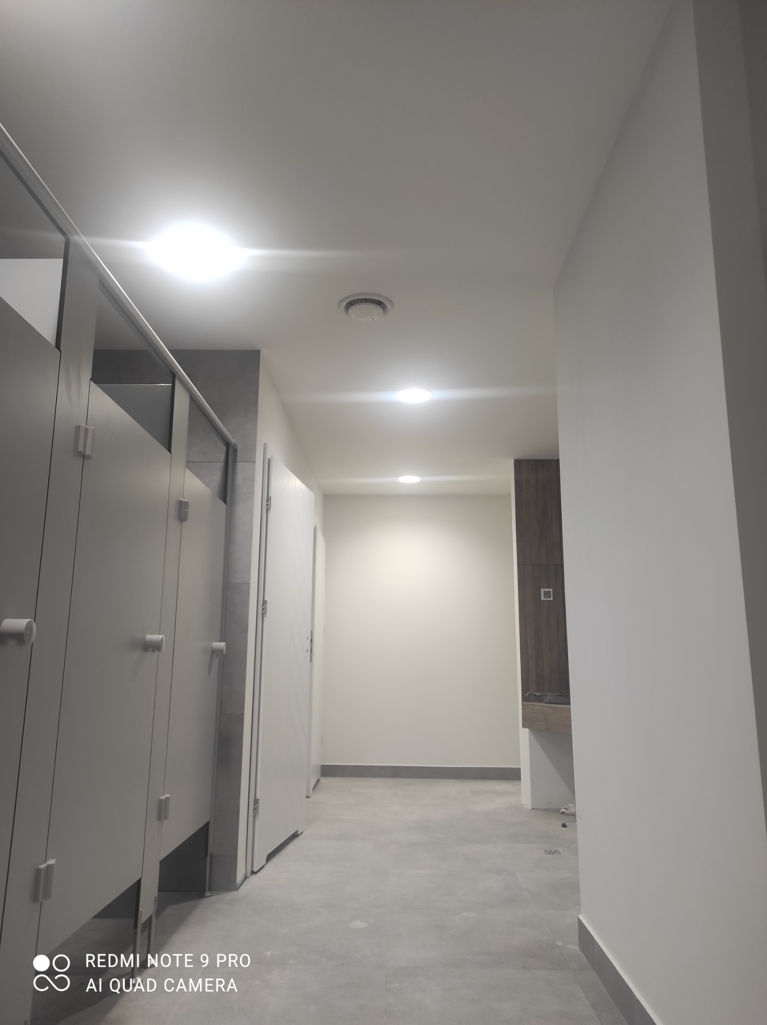 Wnętrze publicznej toalety z kabinami i oświetleniem sufitowym, widok z perspektywy podłogi w kierunku umywalek i szafek w tle.