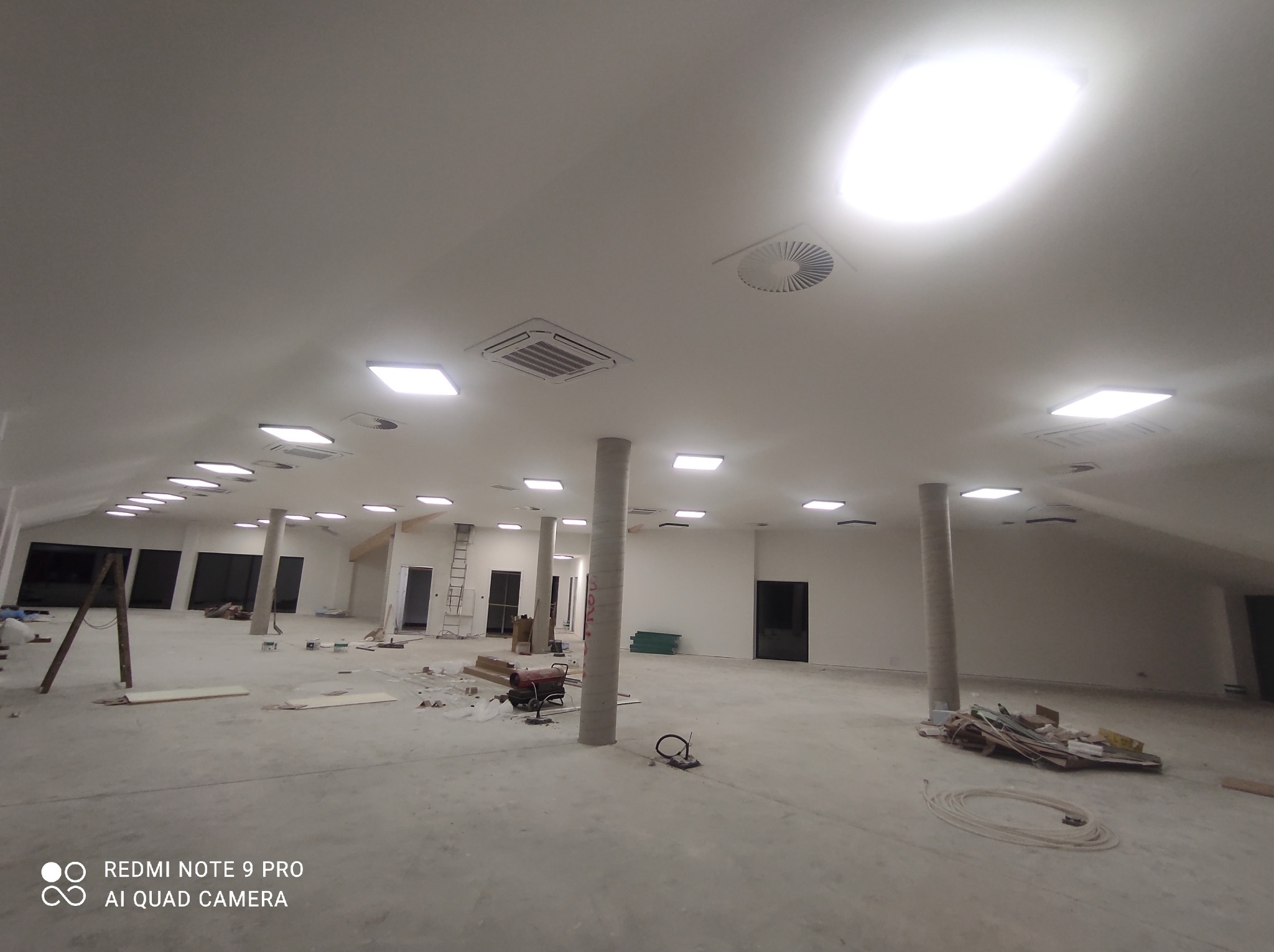 Duża hala w trakcie remontu z widocznymi instalacjami sufitowymi: kwadratowe panele oświetleniowe, okrągłe nawiewniki wentylacyjne i klimatyzacja kasetonowa, na podłodze porozrzucane materiały...