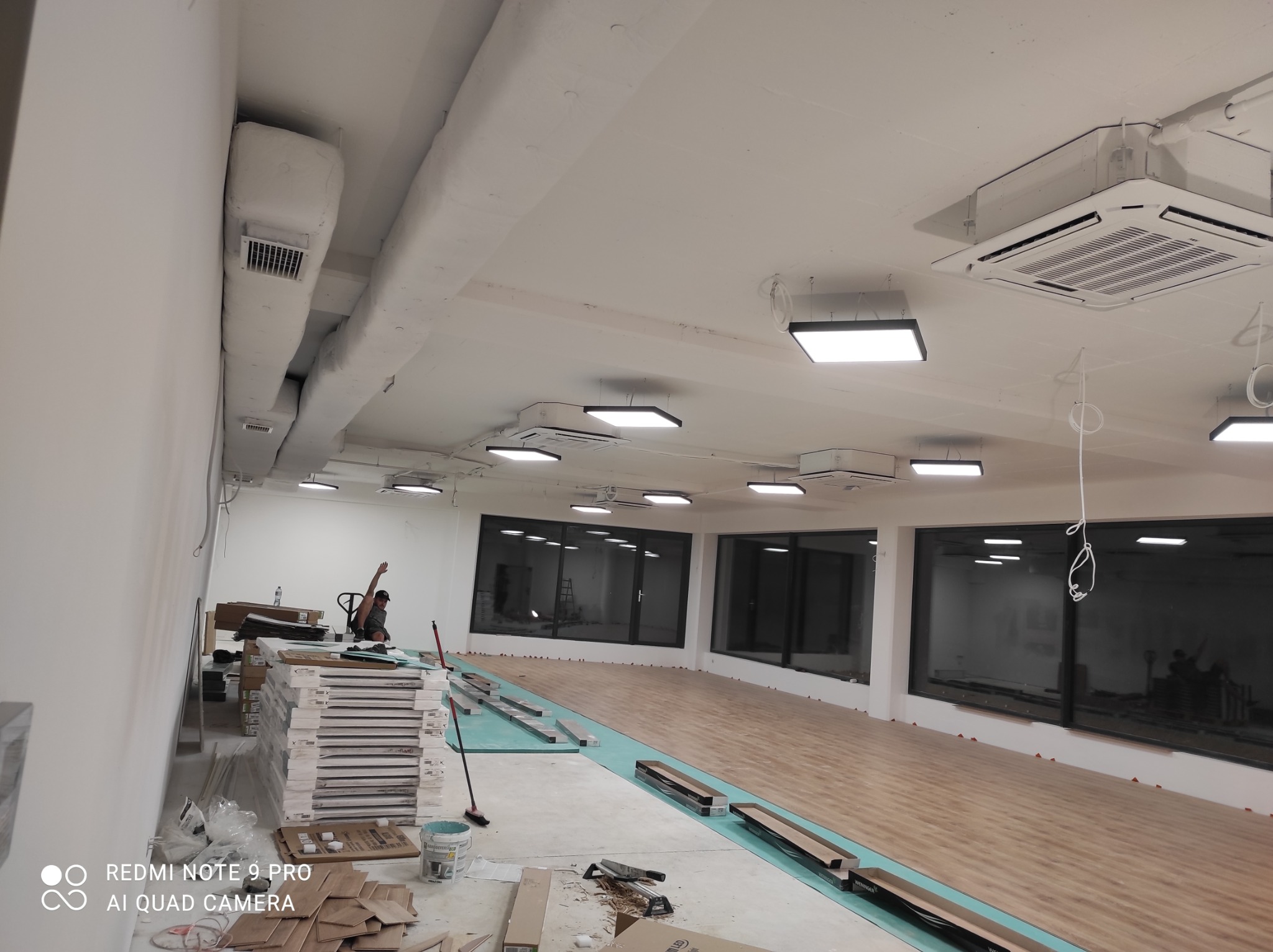 Wnętrze w trakcie montażu oświetlenia i klimatyzacji: widoczne kanały wentylacyjne, klimakonwektory kasetonowe, panele LED i częściowo ułożona podłoga z paneli drewnopodobnych.