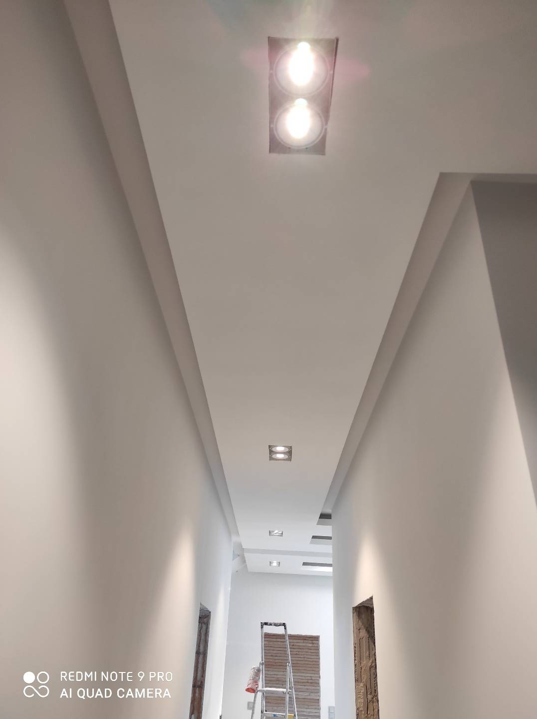 Oświetlenie sufitowe w korytarzu z zabudową gipsowo-kartonową, widoczna drabina i częściowo wykończone ściany.