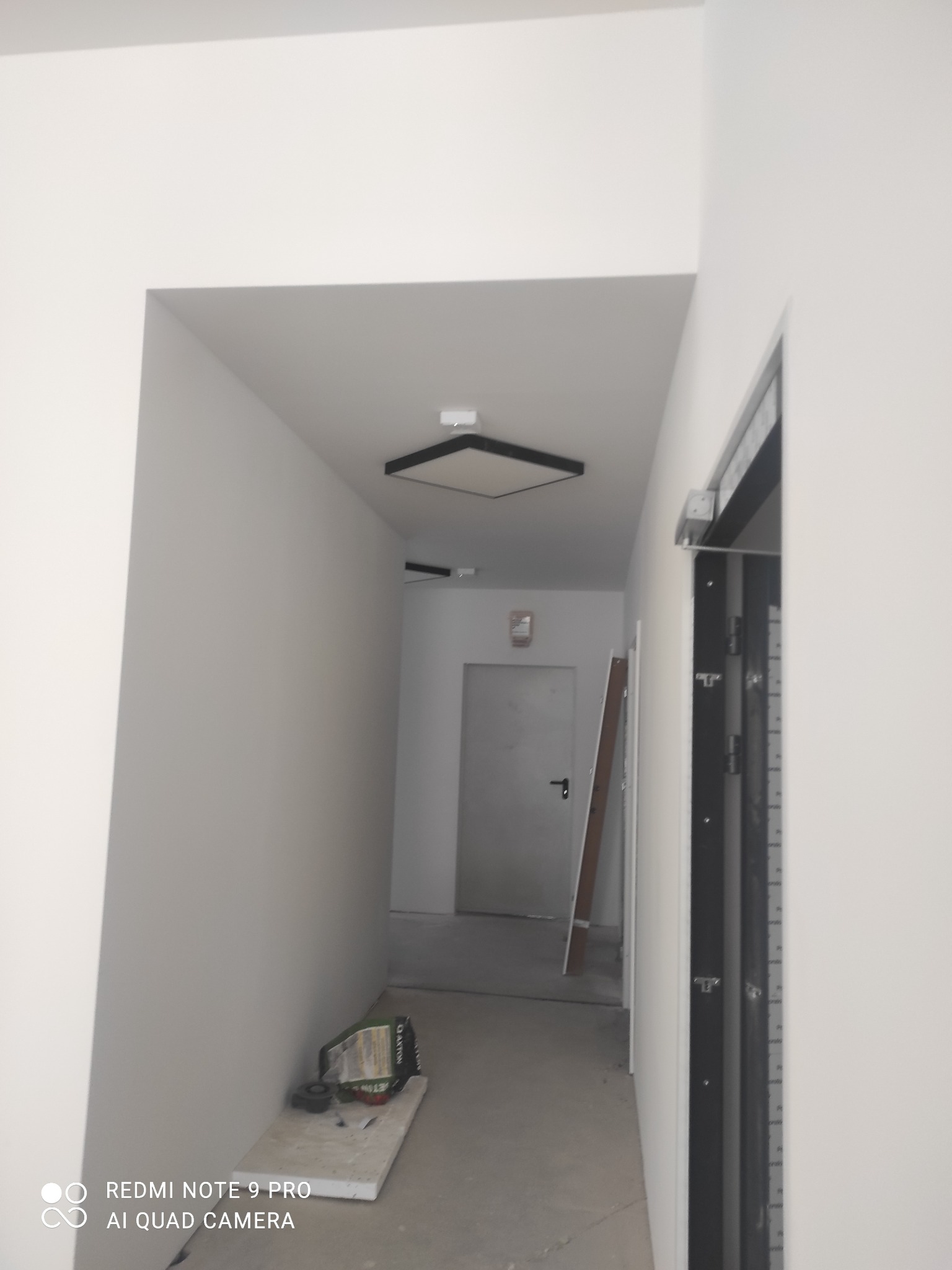 Korytarz w trakcie remontu z zamontowanym kwadratowym panelem LED na suficie, widoczne narzędzia na podłodze i drabina oparta o ścianę w oddali.