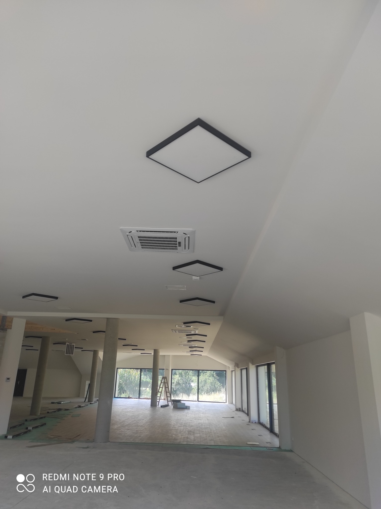 Wnętrze w trakcie wykańczania: widok na sufit z zainstalowanymi kwadratowymi lampami LED z czarną obwódką i klimatyzatorem kasetonowym, widoczne również filary i okna z widokiem na zieleń.