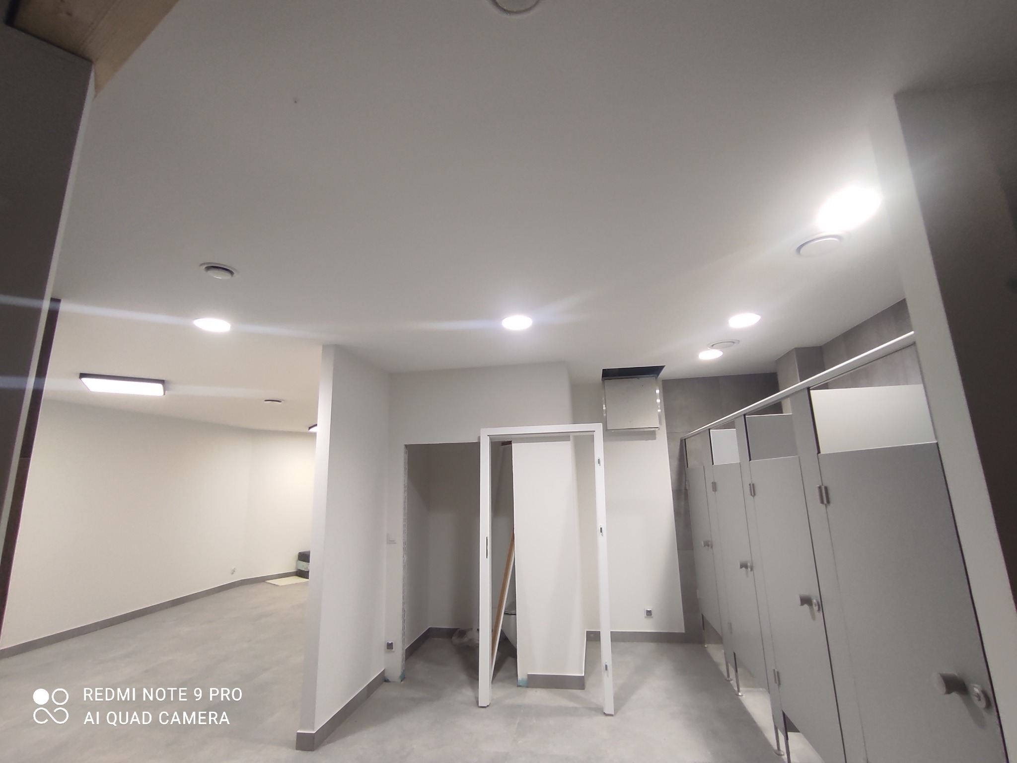 Wnętrze publicznej toalety w trakcie instalacji oświetlenia sufitowego, widoczne kabiny WC i przygotowane otwory na drzwi.