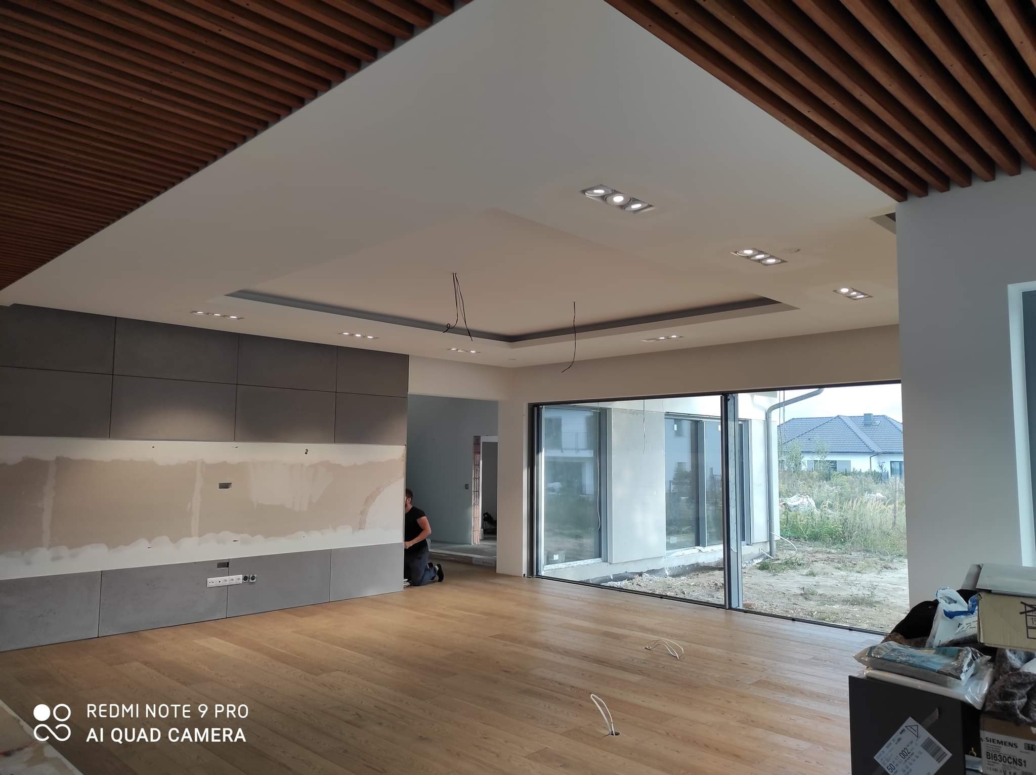 Wnętrze w trakcie wykańczania: drewniana podłoga, szare panele ścienne z widocznymi przewodami elektrycznymi, sufit z drewnianymi listwami i zamontowanymi punktowymi oprawami oświetleniowymi, osoba...