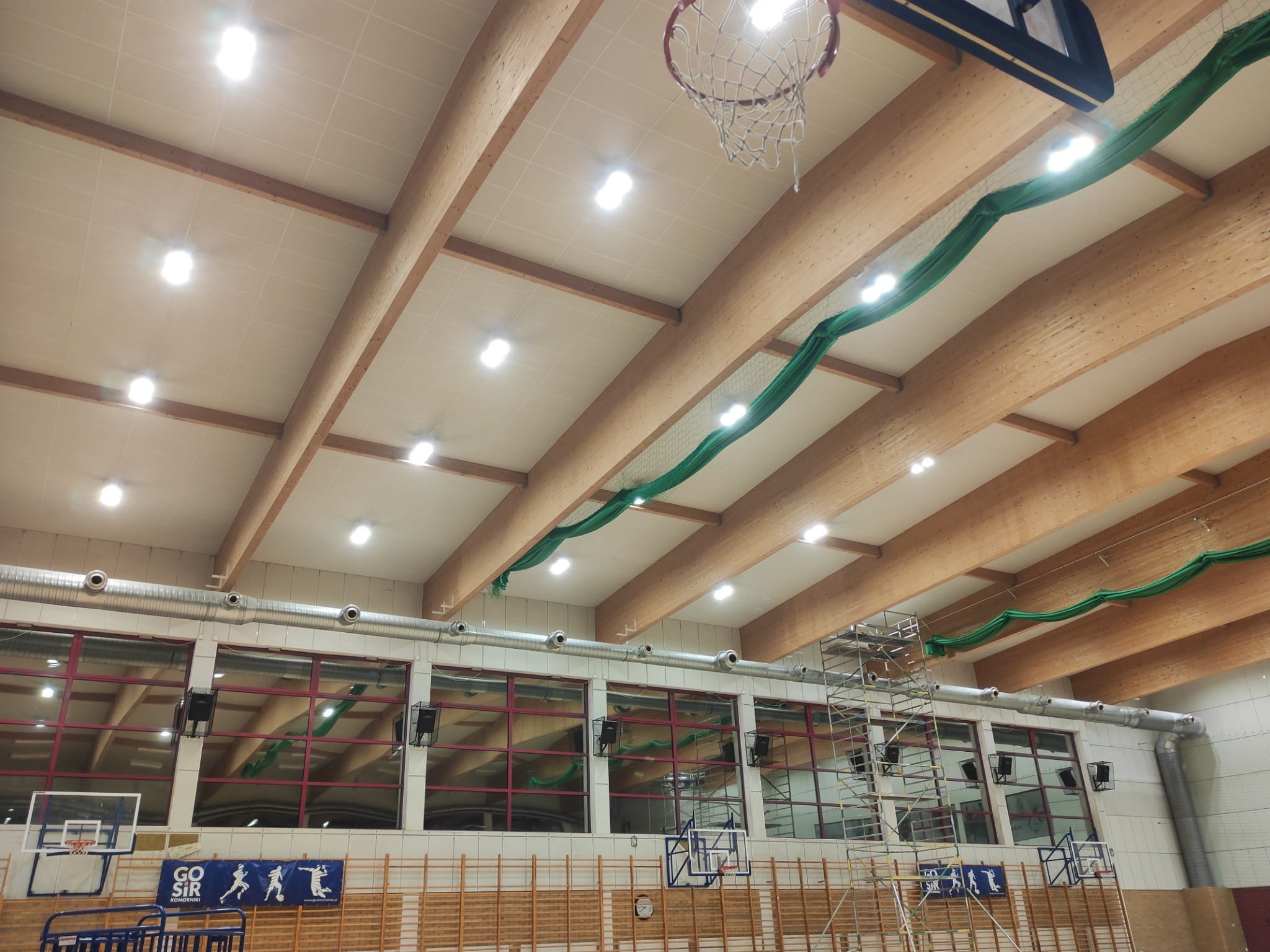 Wysoka hala sportowa z drewnianą konstrukcją dachu i oświetleniem LED, widoczne rusztowania i elementy instalacji wentylacyjnej oraz siatka zabezpieczająca pod sufitem.