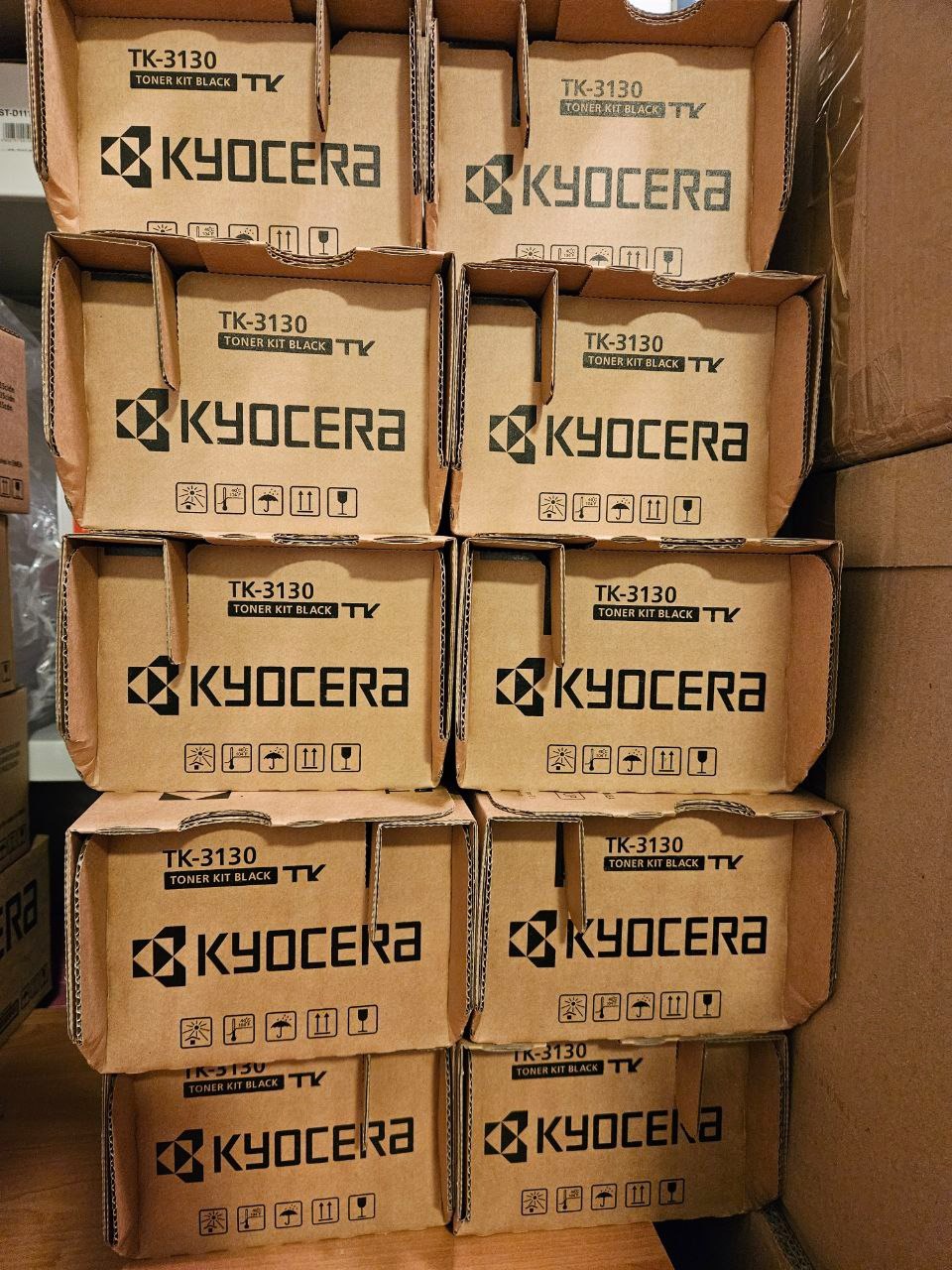 Stos kartonowych pudełek z tonerami Kyocera TK-3130, ułożonych jeden na drugim. Widoczne logo i oznaczenia modelu. Brązowe kartony w magazynowym otoczeniu.