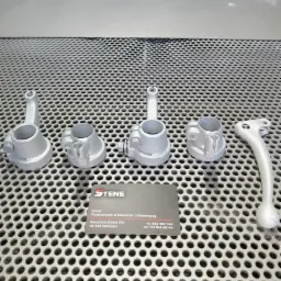 Metalowe elementy po obróbce, ułożone na perforowanym stole warsztatowym, z wizytówką firmy specjalizującej się w obróbce strumieniowo-ściernej w tle i narzędziem pneumatycznym w górnej części kadru.