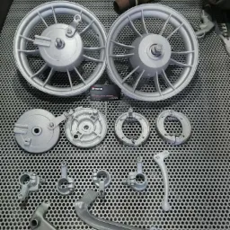 Dwa srebrne koła motocyklowe i inne części, świeżo po obr&oacute;bce, leżą na metalowej kratownicy. Widoczny wąż i pistolet natryskowy w tle.