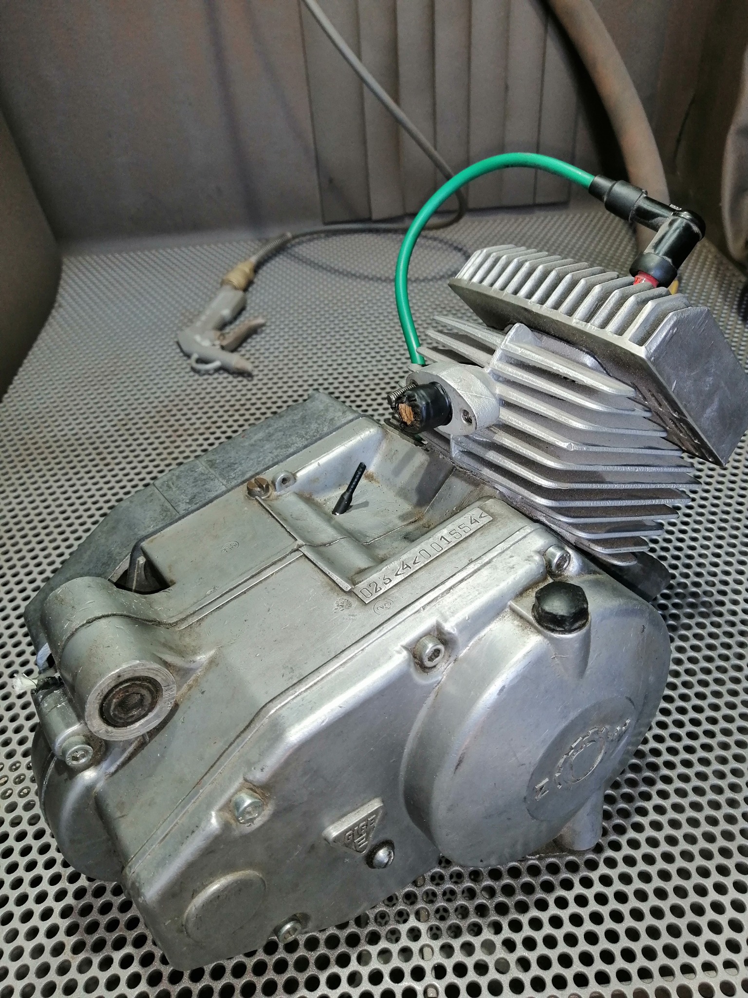 Silnik motocyklowy po czyszczeniu, widoczne ślady korozji i numer seryjny, na tle metalowej kratownicy, obok pistolet do piaskowania.