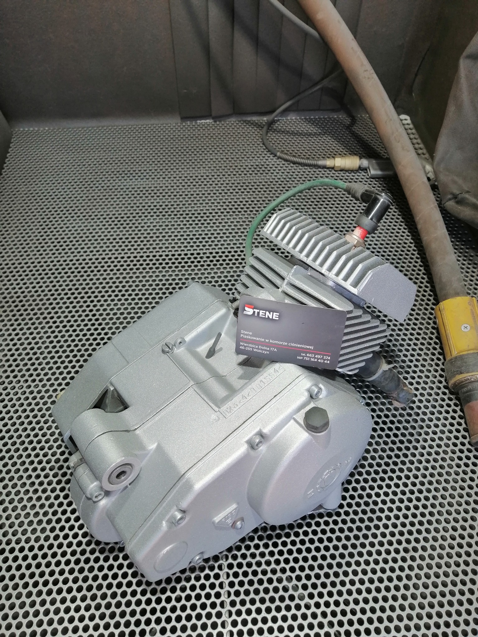 Pomalowany na srebrno silnik motocyklowy leży na perforowanym metalowym stole obok węża i wizytówki firmy oferującej usługi w komorze ciśnieniowej.