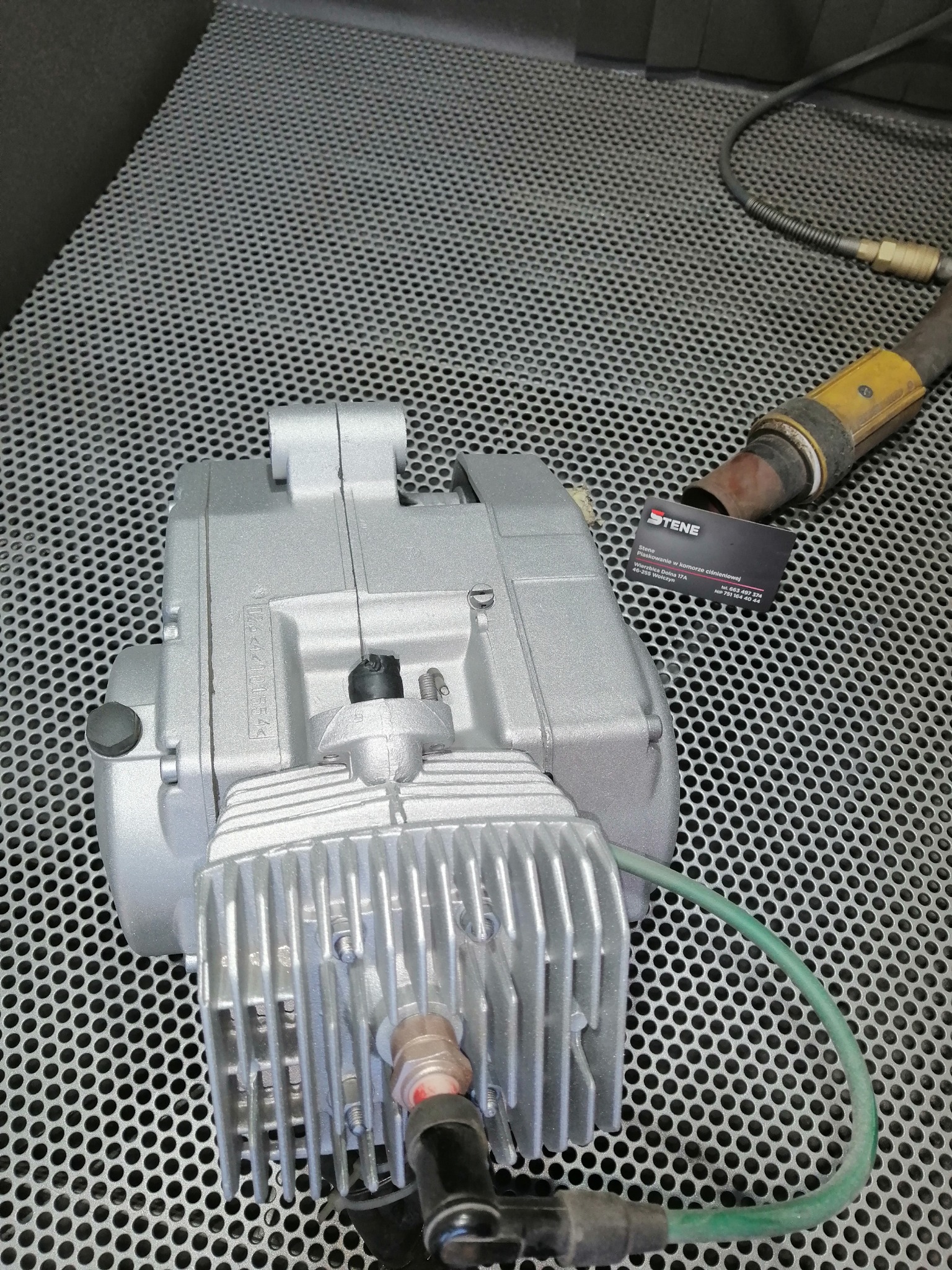 Silnik motocyklowy po obróbce, prawdopodobnie po oczyszczeniu, leżący na perforowanym metalowym stole warsztatowym, obok narzędzia i wizytówki firmy oferującej usługi w komorze ciśnieniowej.