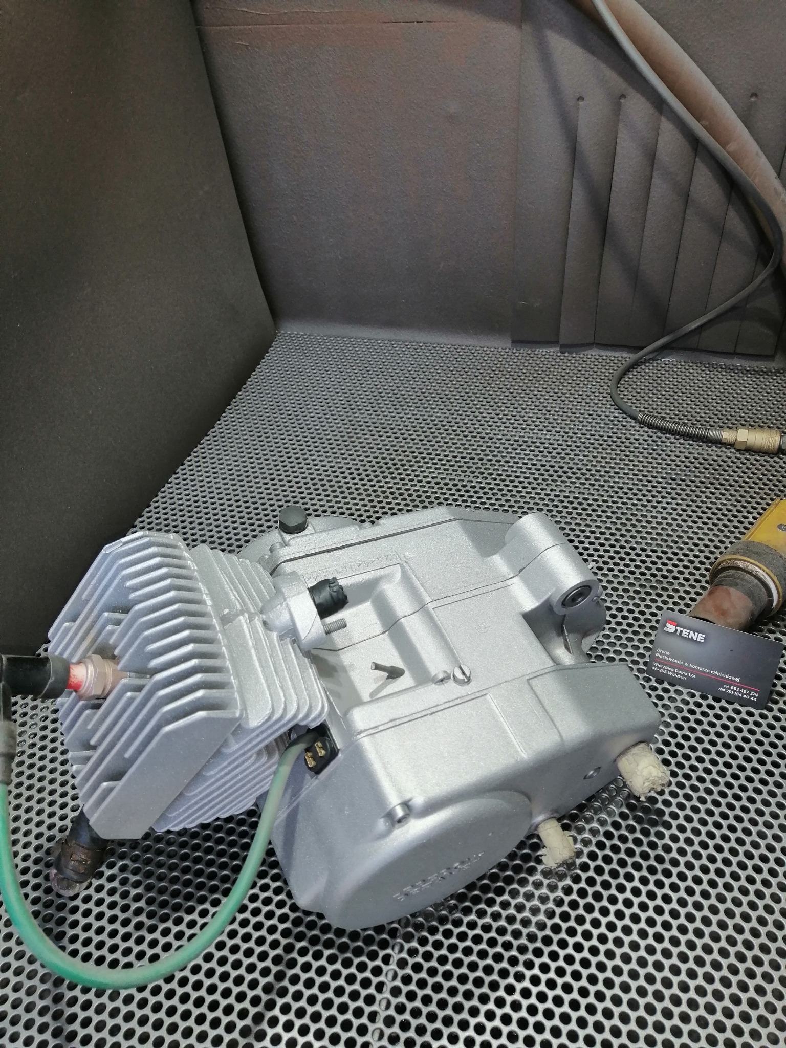 Szczegółowy widok silnika motocyklowego po renowacji, z świeżo nałożoną, srebrną powłoką ochronną, umieszczonego na perforowanym metalowym stole w warsztacie. W tle widoczna ściana kabiny...