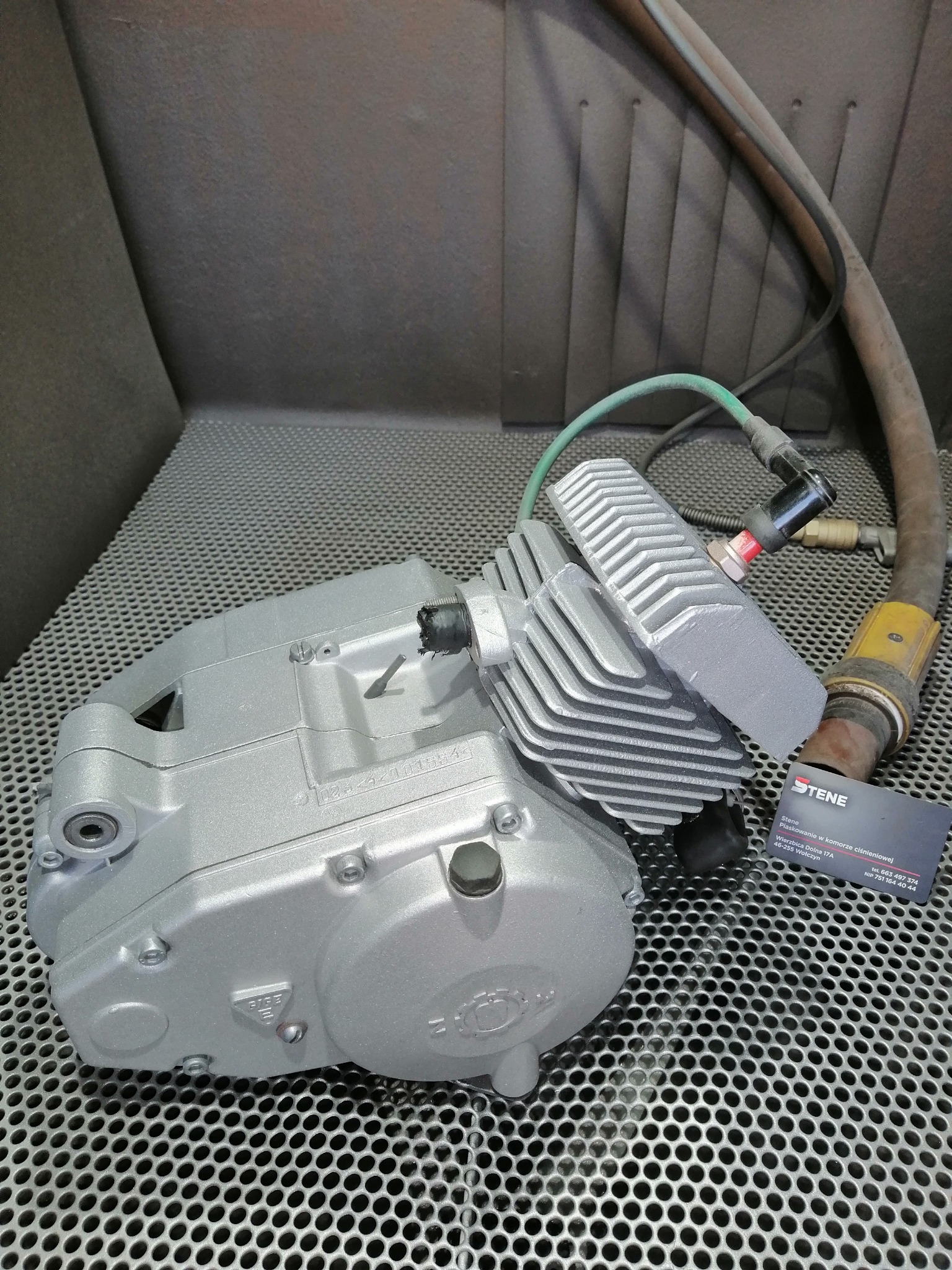 Silnik motocyklowy po obróbce w komorze, świeżo pokryty jednolitą, srebrną powłoką, leżący na metalowej kratownicy. Widoczny przewód i wizytówka firmy.