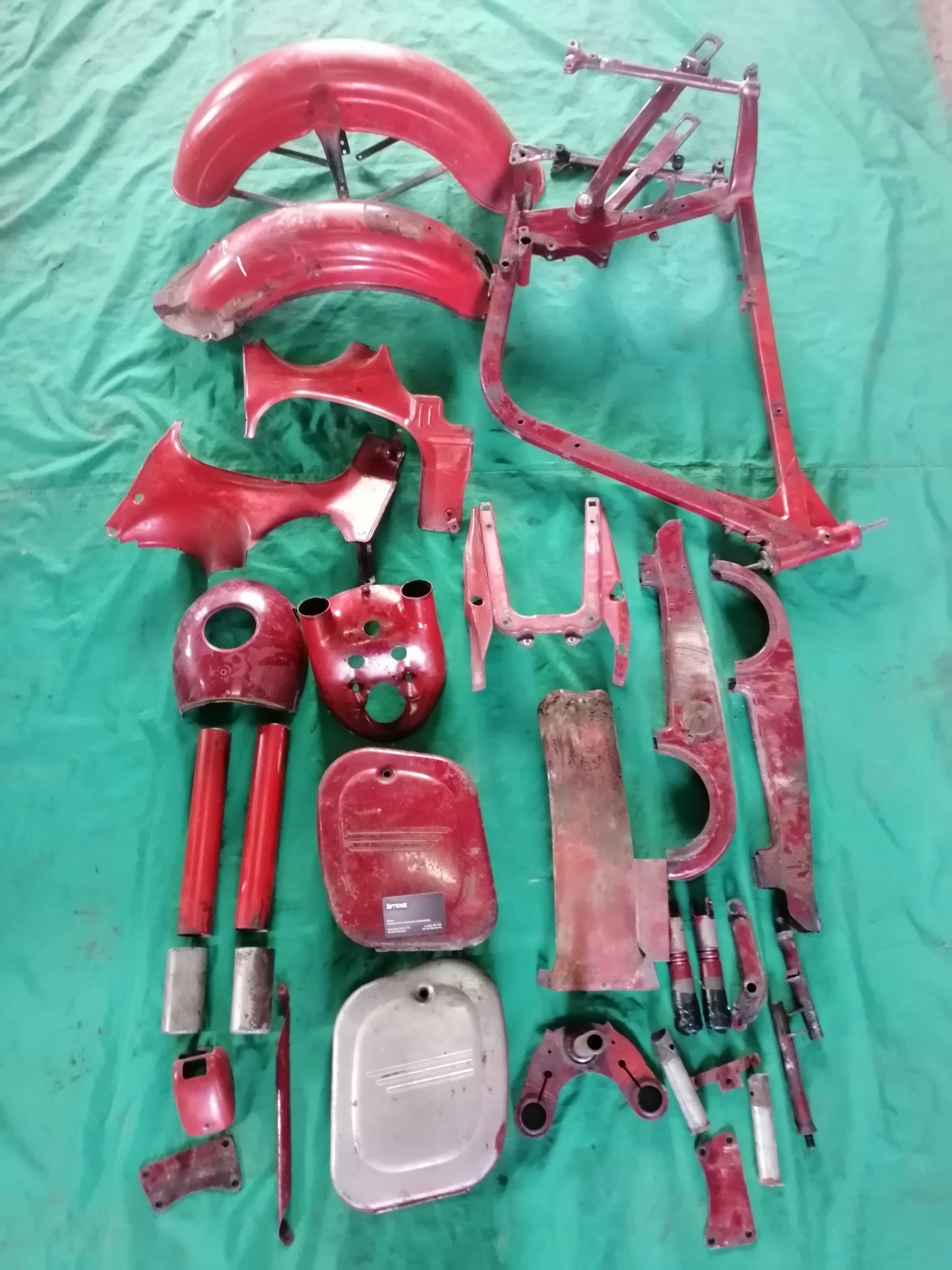 Części motocyklowe po usunięciu starej farby, przygotowane do dalszej obróbki, widoczna rama, błotniki i inne elementy w kolorze czerwonym na zielonym tle.