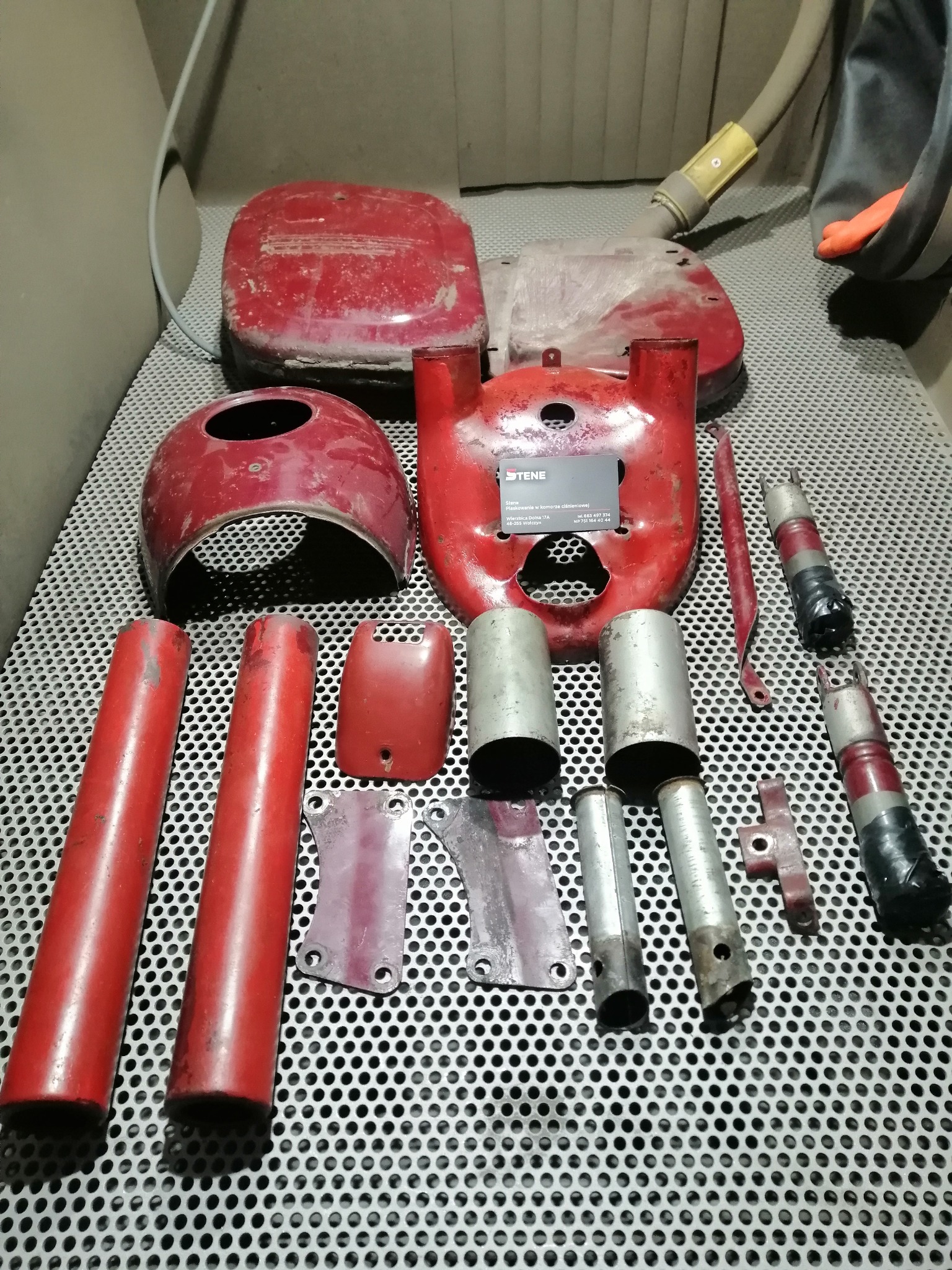 Elementy motocyklowe, niektóre w czerwonej farbie, inne w kolorze metalu, ułożone na perforowanym stole w komorze do obróbki strumieniowo-ściernej, widoczny wąż odciągowy i rękawica ochronna.