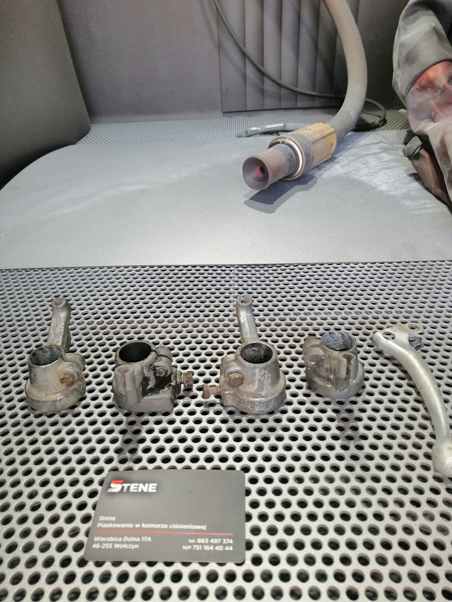 Metalowe elementy motocyklowe przed obróbką, leżące na perforowanym stole w komorze, widoczny wąż do piaskowania i wizytówka firmy Stene specjalizującej się w piaskowaniu.