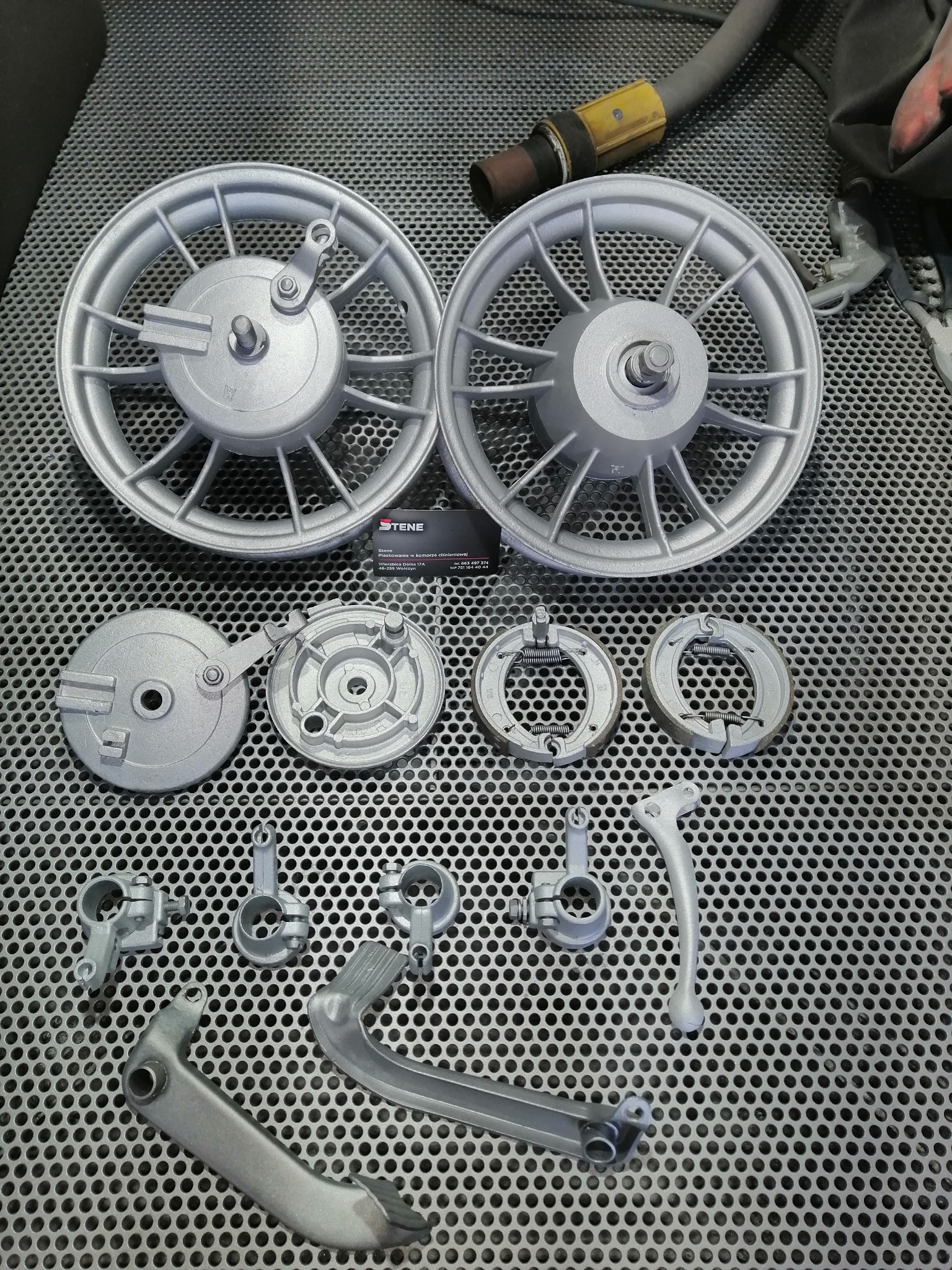 Dwa srebrne koła motocyklowe i inne części, świeżo po obróbce, leżą na metalowej kratownicy. Widoczny wąż i pistolet natryskowy w tle.