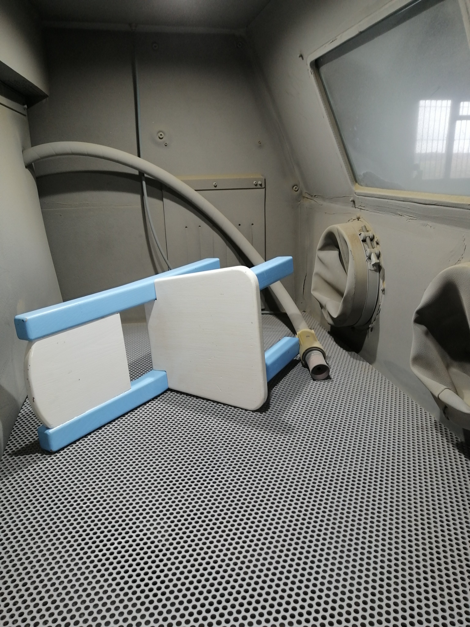 Biało-błękitne krzesełko dziecięce w komorze do piaskowania, widoczny przewód od odkurzacza przemysłowego i metalowa kratownica podłoża.