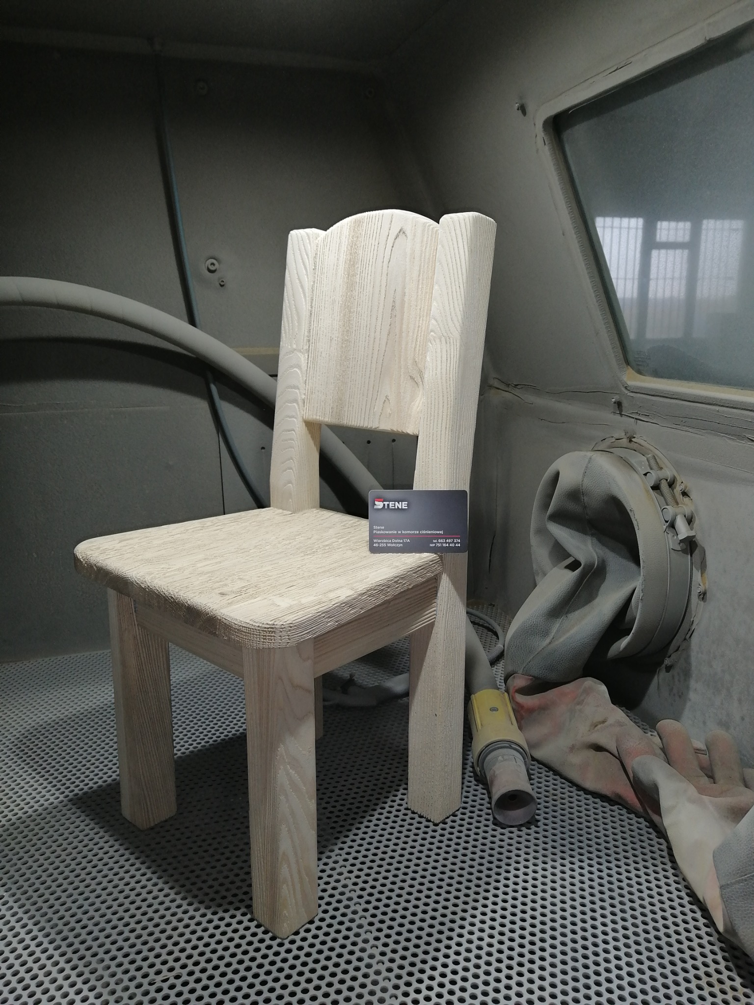Drewniane krzesło dziecięce po obróbce w komorze, leży na nim wizytówka firmy zajmującej się obróbką strumieniowo-ścierną. Obok leży rękawica ochronna i element instalacji.