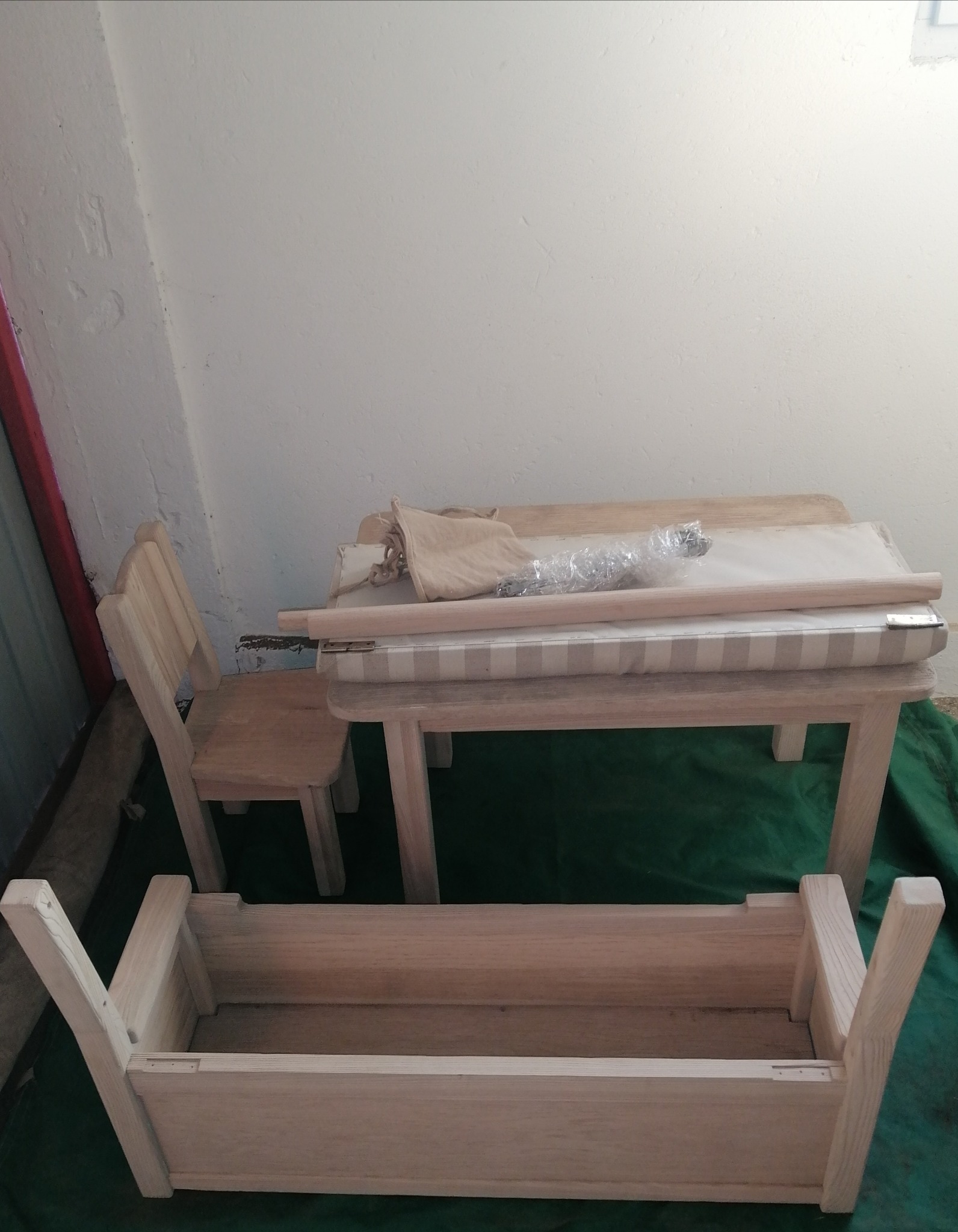 Niedokończony zestaw mebli dziecięcych z surowego drewna: stół z tapicerowanym blatem, krzesło oraz ławka z odwróconymi nogami, na zielonym tle.