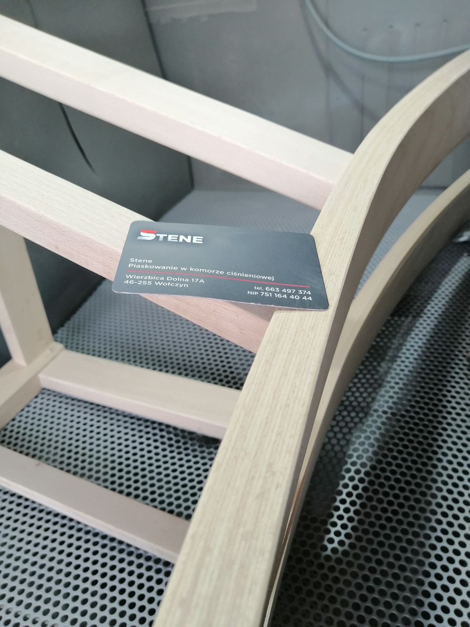 Drewniany szkielet krzesła w komorze, z wizytówką firmy oferującej usługi obróbki strumieniowo-ściernej leżącą na elemencie konstrukcyjnym.