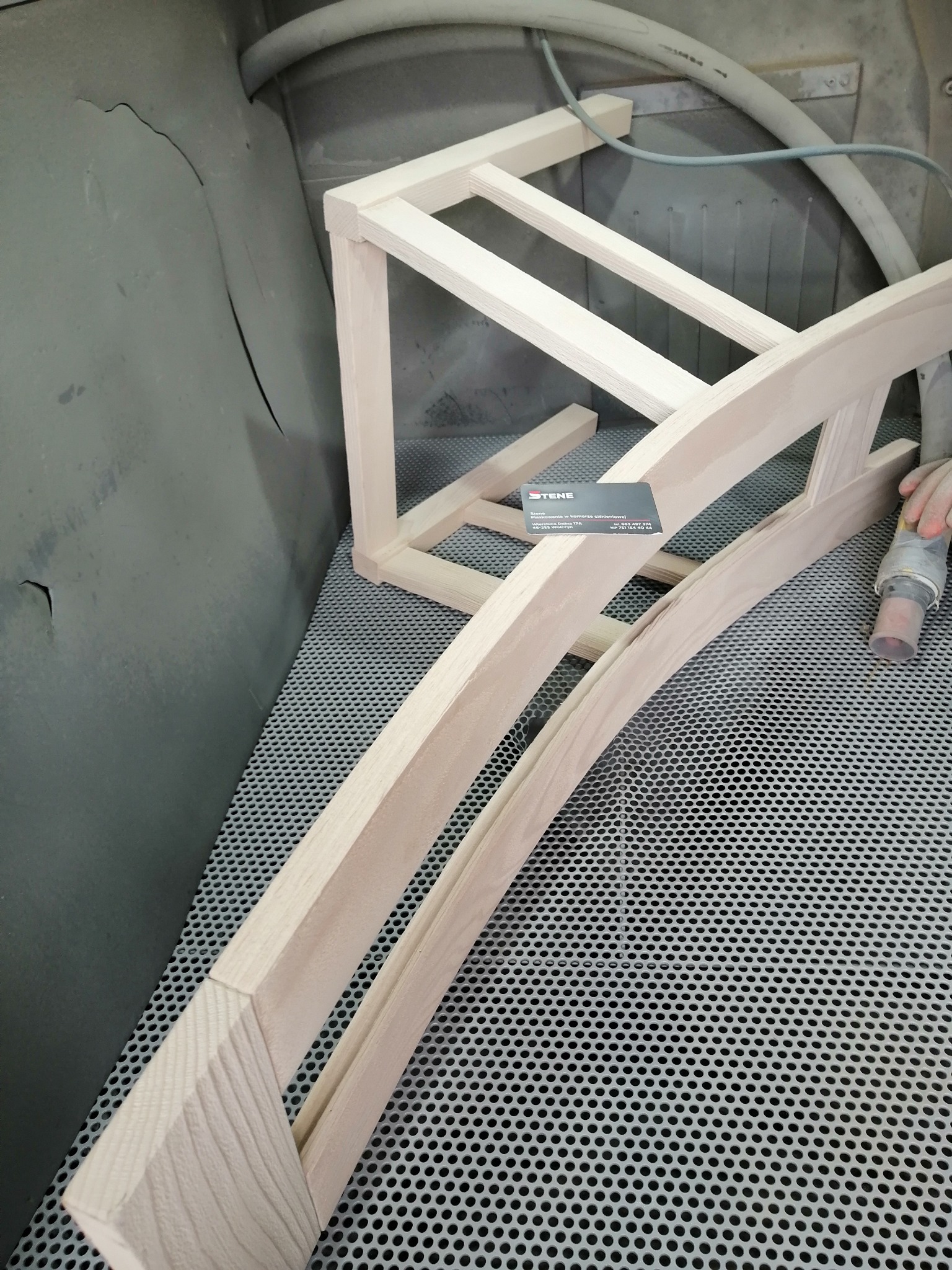 Elementy drewniane krzesła w komorze do obróbki powierzchni, widoczna dysza pistoletu roboczego i wizytówka firmy.