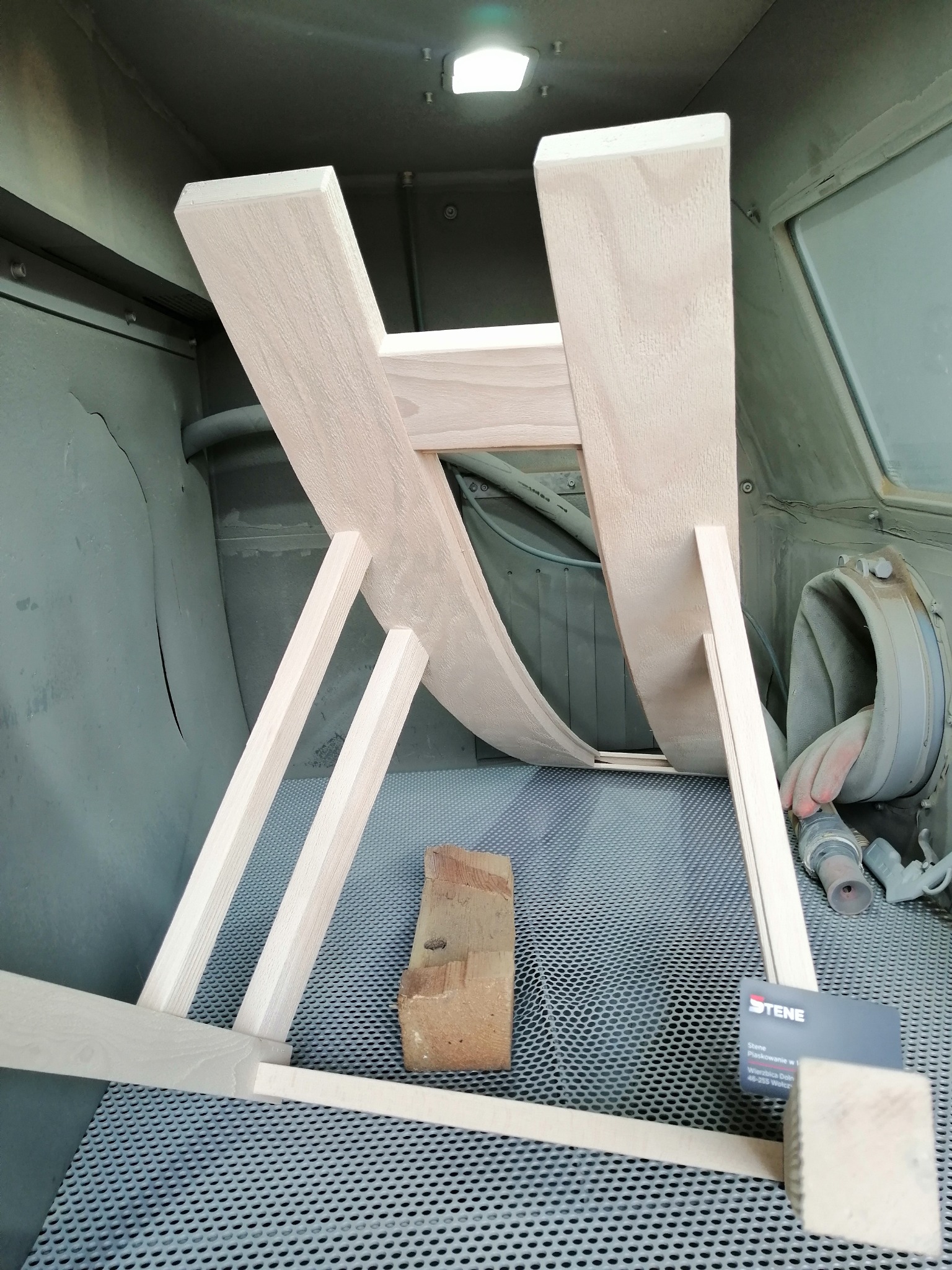 Drewniany stelaż krzesła wewnątrz kabiny lakierniczej, wsparty na cegle, przed procesem wykończenia, widoczny system wentylacyjny i wizytówka firmy.