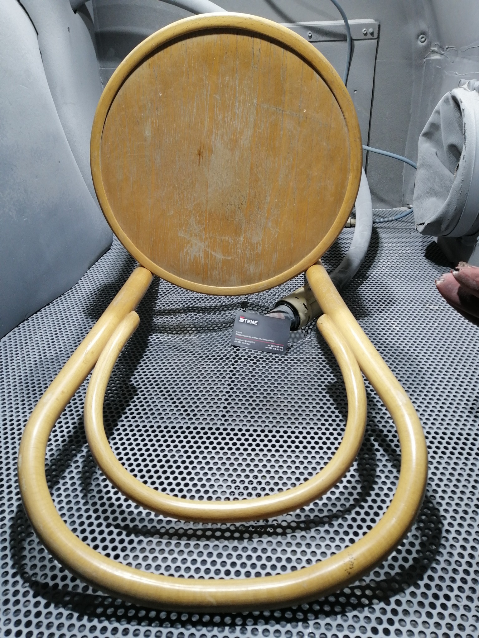Krzesło Thonet przed piaskowaniem w komorze, widoczne ślady użytkowania na siedzisku, metalowa podłoga komory piaskarskiej.