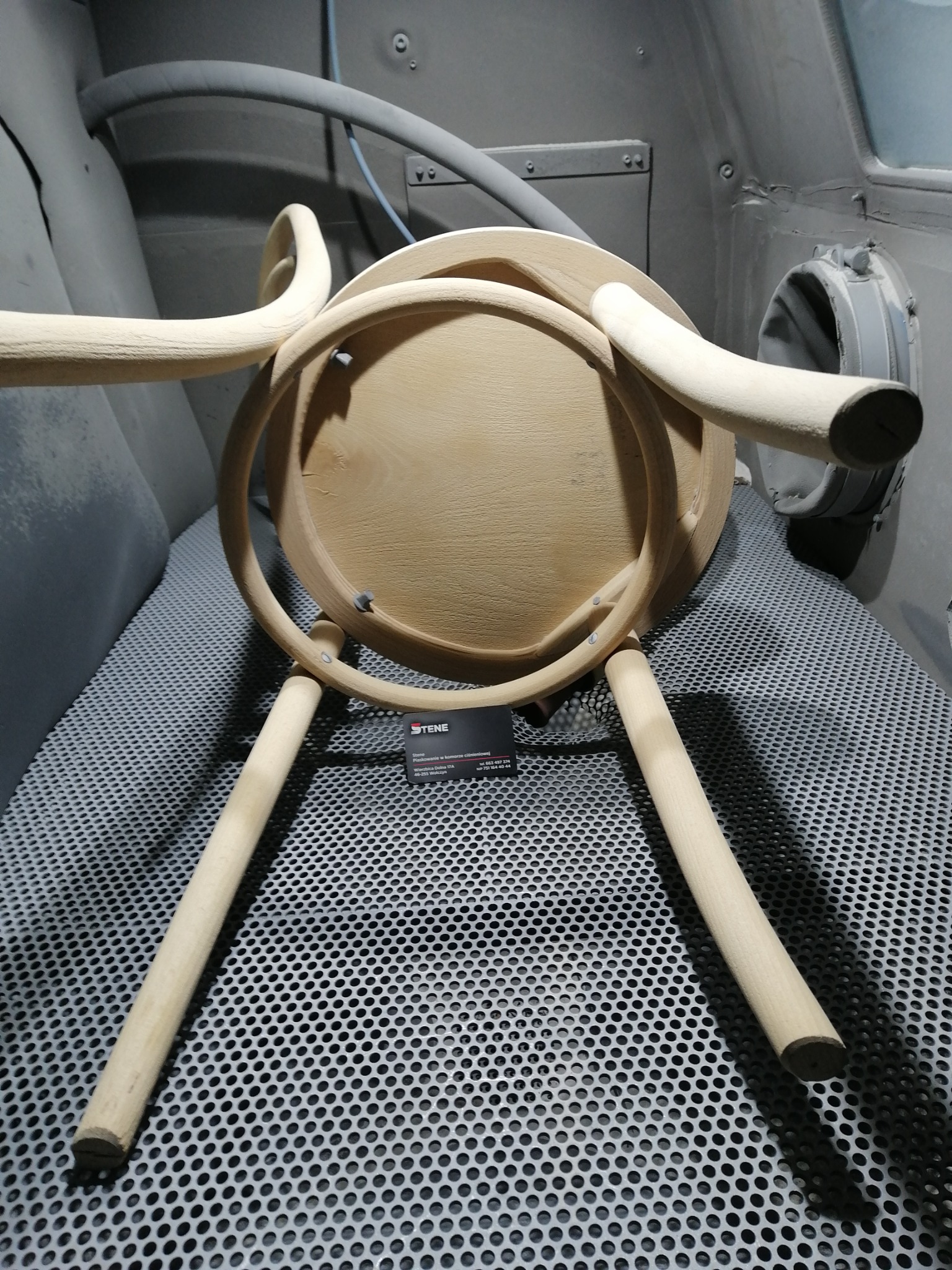 Drewniane krzesło Thonet przed obróbką, widoczne ślady po procesie czyszczenia, umieszczone na perforowanej podłodze w szarej komorze.