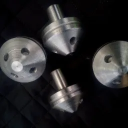 Cztery metalowe elementy z otworami i stożkowymi zakończeniami, ułożone na czarnym, pikowanym tle. Widoczne ślady obróbki skrawaniem.