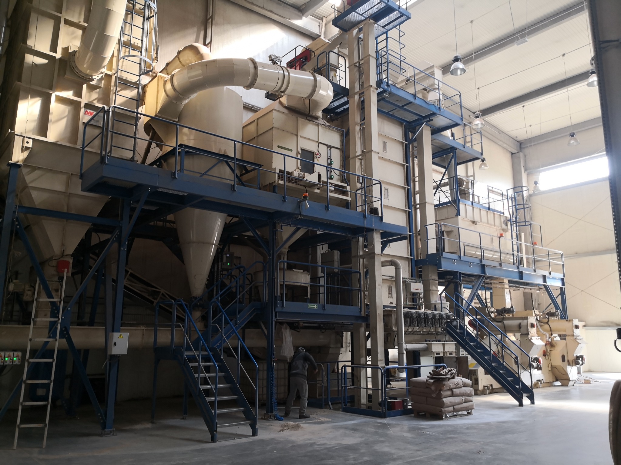 Wnętrze hali produkcyjnej z dużą instalacją przemysłową do przetwarzania biomasy, widoczne rury, schody, platformy, worki z gotowym produktem oraz sylwetka pracownika w tle.