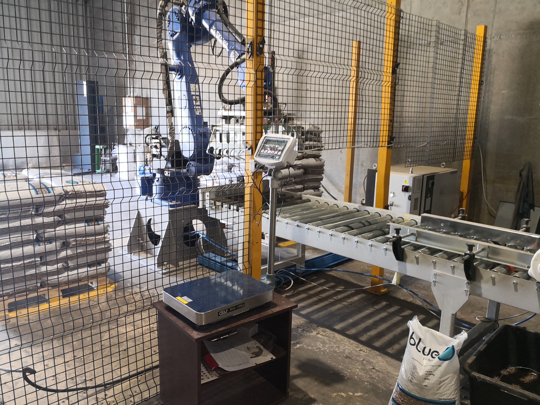 Zautomatyzowana linia produkcyjna z robotem przemysłowym pakującym worki z pelletem, widoczna waga i przenośnik rolkowy, całość za siatką ochronną.