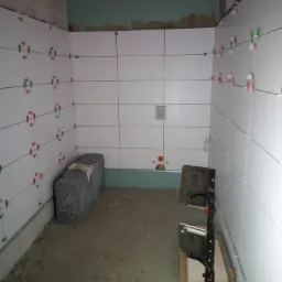 Ściany w trakcie układania białych płytek w łazience, widoczne krzyżyki dystansowe, fragment instalacji hydraulicznej oraz narzędzia glazurnicze na podłodze.