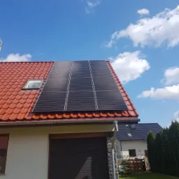 Panele słoneczne na czerwonym dachu domu jednorodzinnego z oknem dachowym, błękitne niebo z obłokami w tle.